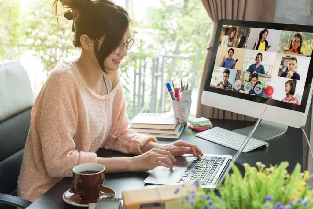 A woman works from home at a laptop on a spacious desk while a virtual meeting appears on another monitor