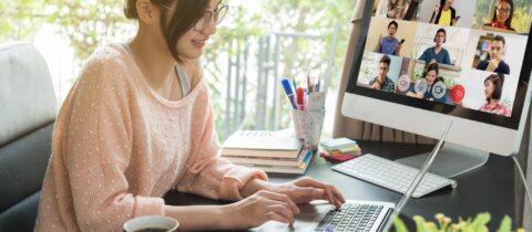 A woman works from home at a laptop on a spacious desk while a virtual meeting appears on another monitor