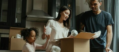 A family of three packs items in a kitchen, using cardboard boxes and bubble wrap.