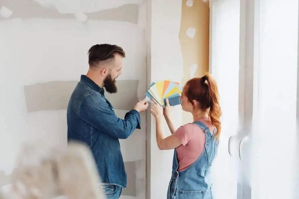 A couple holds up paint swatches to a blank wall as they decide what color is best