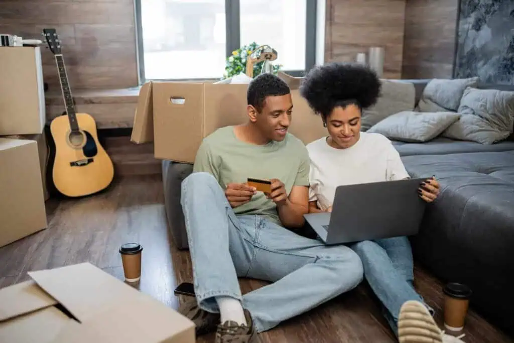 A couple sitting on the floor surrounded by moving boxes looks at a laptop. One of the pair is holding a credit card.