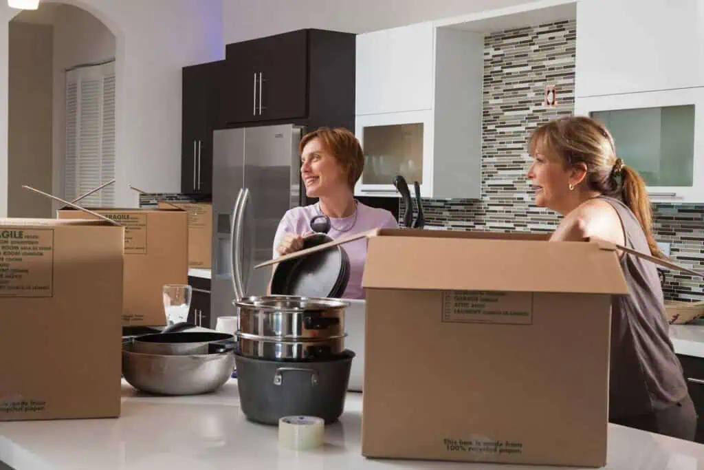 Two women lean against a kitchen countertop. There are several boxes as well as pots and pans on the counter as well. 