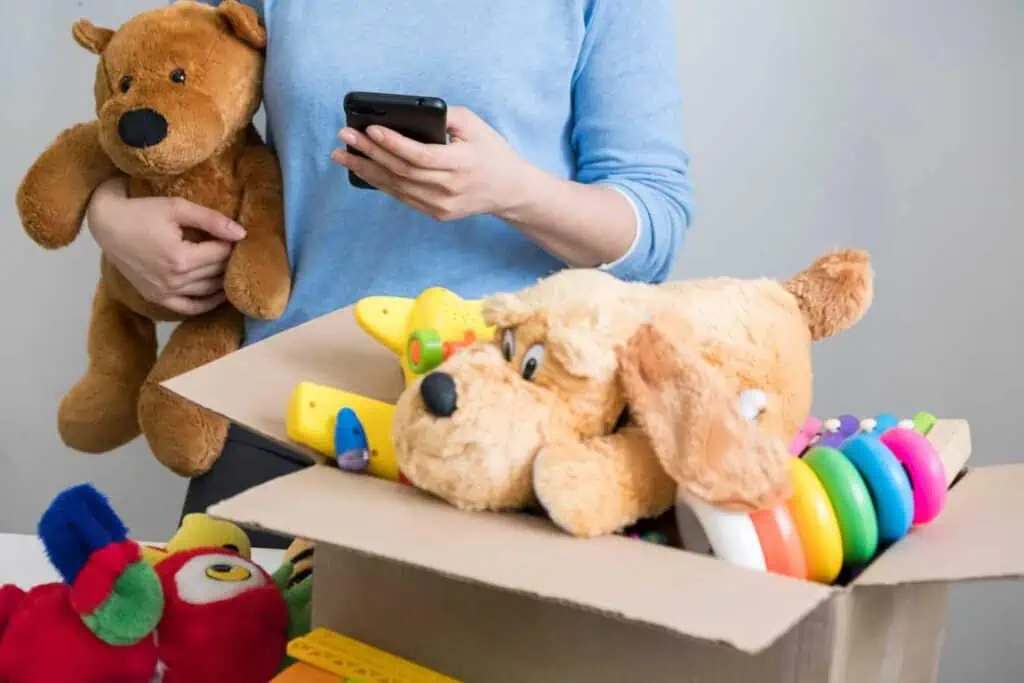 A person looks something up on their phone. A box of toys is set in front of them and they have a teddy bear under the crook of their arm