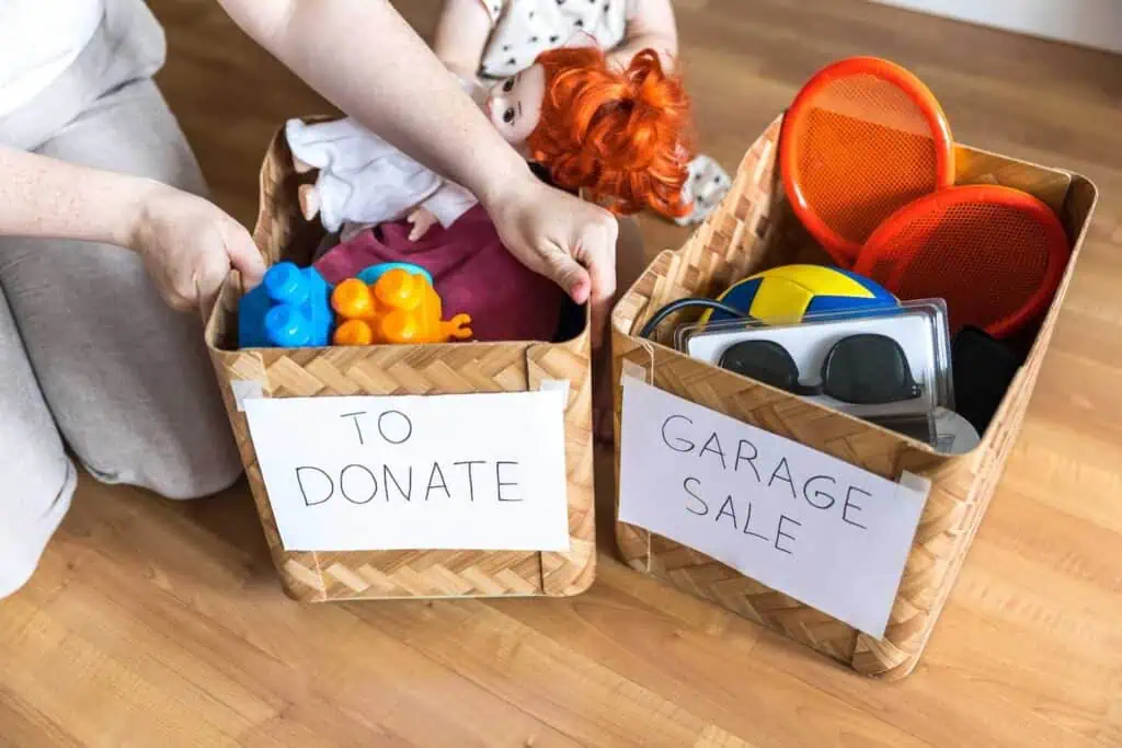 A person grips the edge of a box with a sign labeled "to donate" on it. Next to it is another box labeled "Garage sale". Both are full of toys