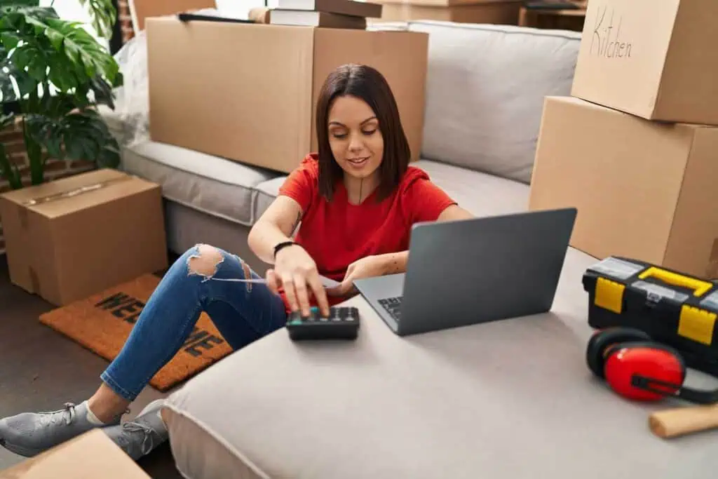 A woman sits on the floor and inputs something on a calculator and her laptop. There are moving boxes around her
