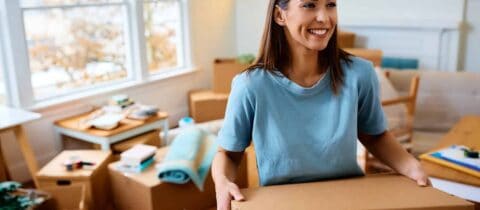 A woman happily carries a cardboard moving box. There are several more behind her in the room she&#039;s in.