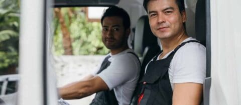 two movers in work overalls sit in the bed of a truck and look at the camera