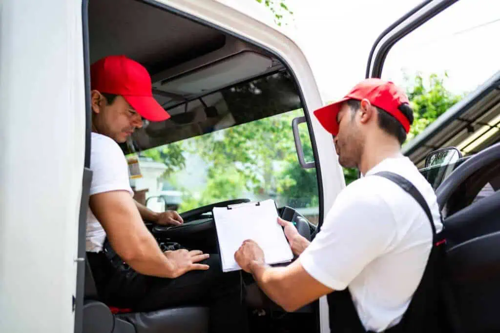 One mover in a red hat discusses something on a clipboard with his colleague sitting in the bed of a truck