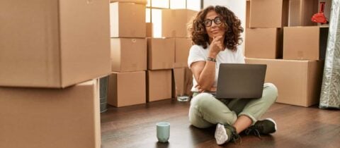 a woman sits on the floor with a laptop and coffee mug. She is surrounded by moving boxes and look contemplative