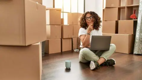 a woman sits on the floor with a laptop and coffee mug. She is surrounded by moving boxes and look contemplative