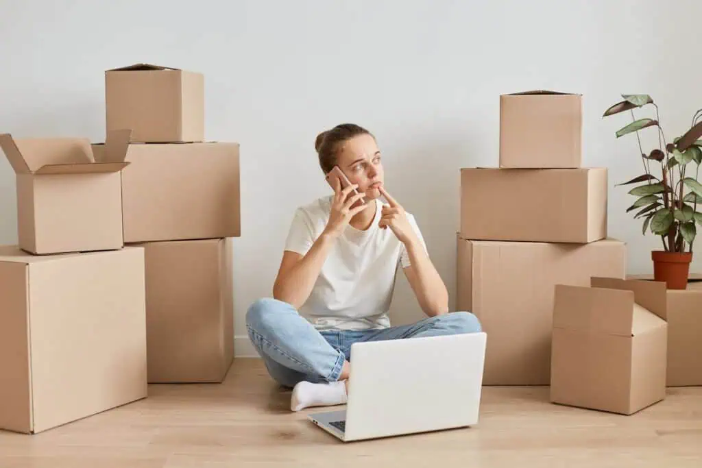 A woman sits contemplatively on the floor with stacks of moving boxes around her. She's speaking on the phone and there is a lap top in front of her