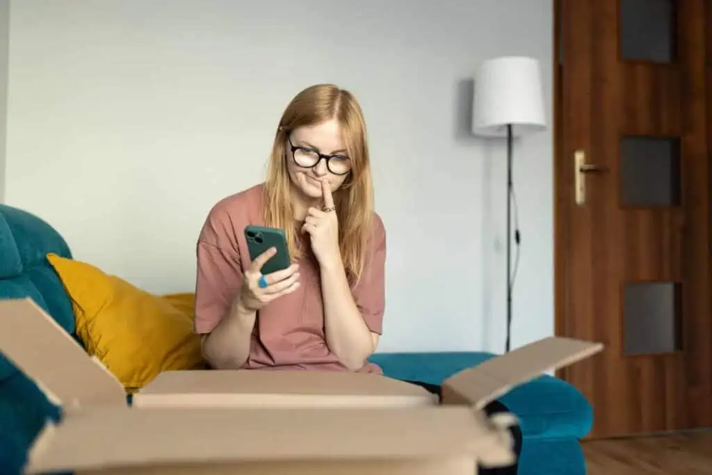 A woman sits on a chair in front of an open moving box. She has a contemplative expression and is holding her phone in one hand. 