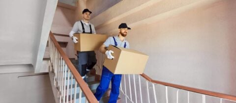 Movers in blue overalls carry boxes down a set of stairs in an apartment building