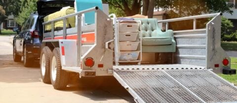 a car with a moving trailer hitched to it sits in a driveway. the trailer is full of furniture.