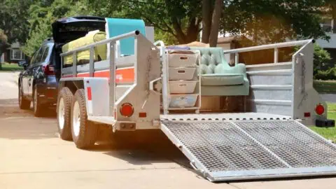 a car with a moving trailer hitched to it sits in a driveway. the trailer is full of furniture.
