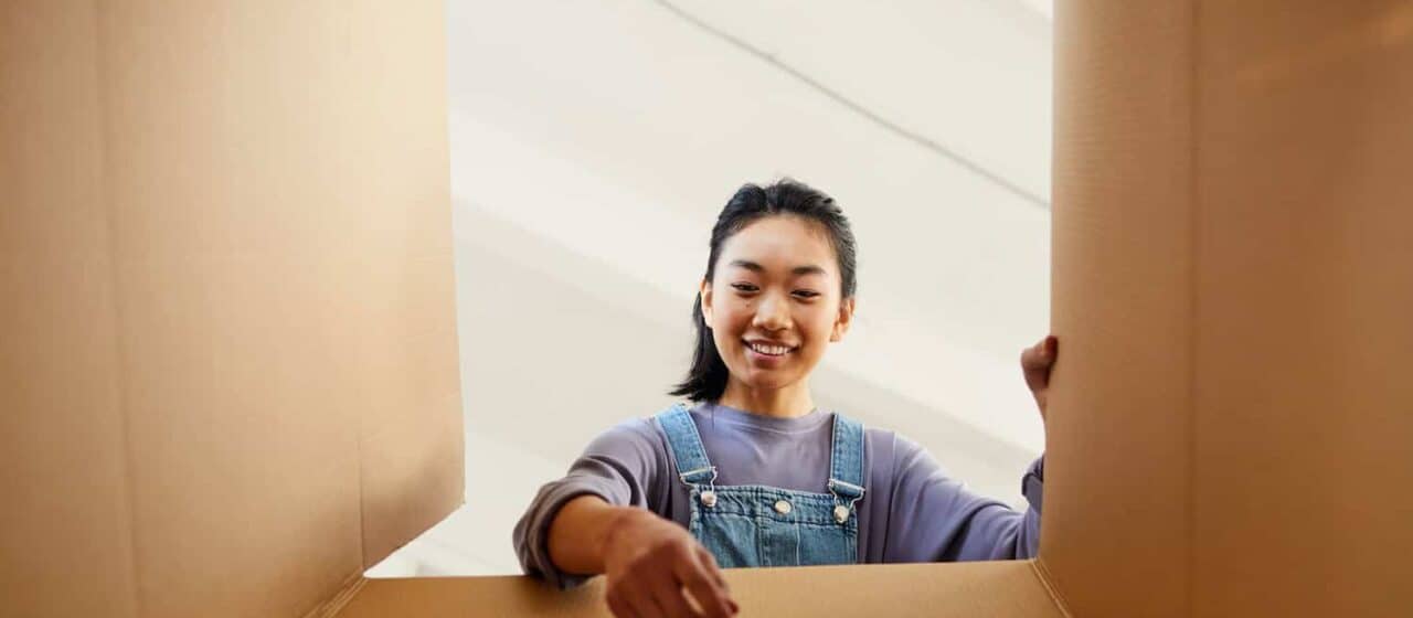 A woman sets down an item into a box. The angle of the shot is from the bottom of the box pointing upwards, so it looks as through the woman is reaching towards the camera