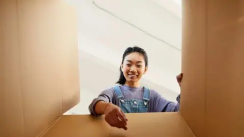 A woman sets down an item into a box. The angle of the shot is from the bottom of the box pointing upwards, so it looks as through the woman is reaching towards the camera