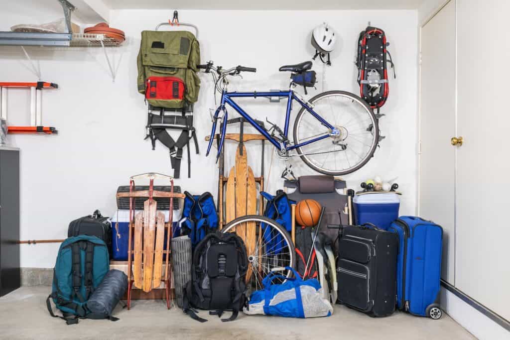 Bikes, sports equipment, several backpacks, a cooler, and sleds are all set against or hung on a garage wall