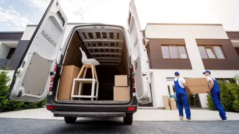 movers carry a box into a home. There are more items in the back of an open moving truck