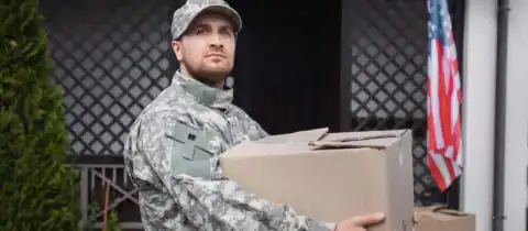 Soldier holding a moving box in front of a house with an American flag.