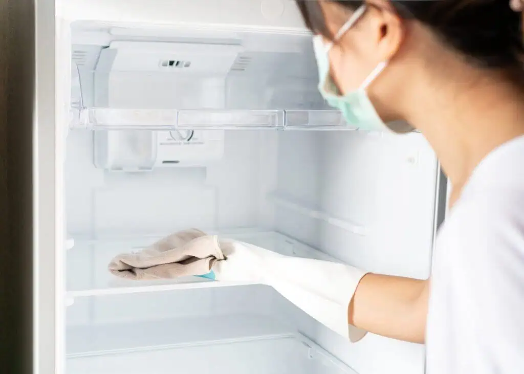 A woman wearing a mask and gloves cleans the inside of a fridge with a towel
