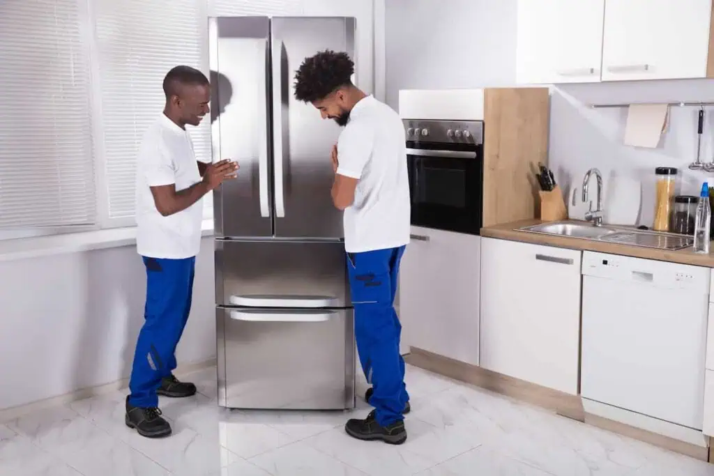 two men in white shirts and blue pants stand on either side of a fridge in a kitchen
