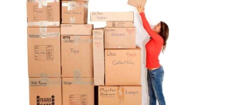 A woman reaches up to place a box at the top of a large stack of them, all labeled with various rooms and categories