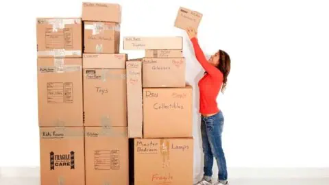 A woman reaches up to place a box at the top of a large stack of them, all labeled with various rooms and categories