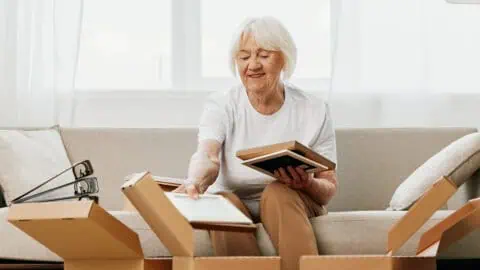 Elderly woman sits on a sofa at home with boxes collecting things with memories albums with photos and photo frames moving to a new place cleaning things and a happy smile.
