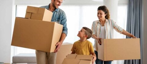 A father, mother, and young son all happily carry packed moving boxes