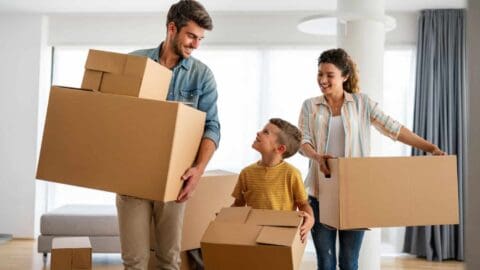 A father, mother, and young son all happily carry packed moving boxes