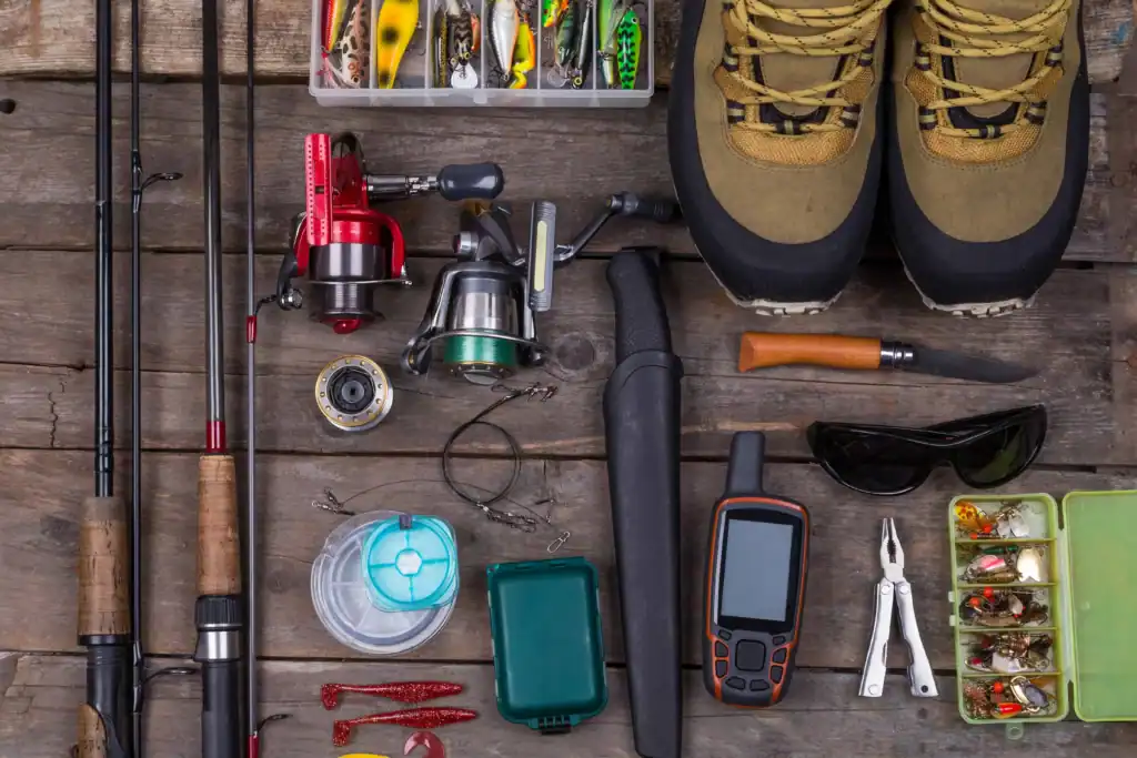 Assorted fishing equipment including rods, reels, lures, boots, and tools arranged on a wooden surface.
