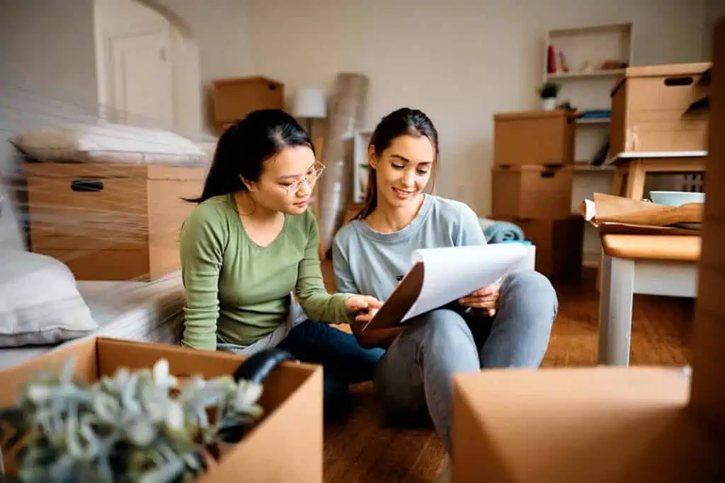 two young women look over a clipboard while they sit in a living room filled with packed boxes