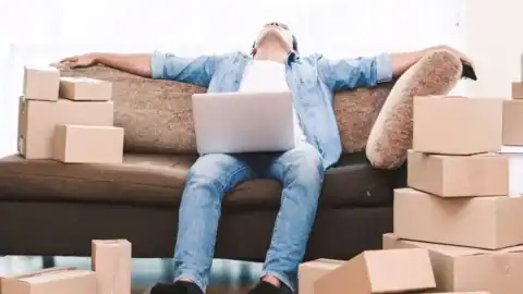 Man overwhelmed by moving stress, surrounded by cardboard boxes and laptop on couch