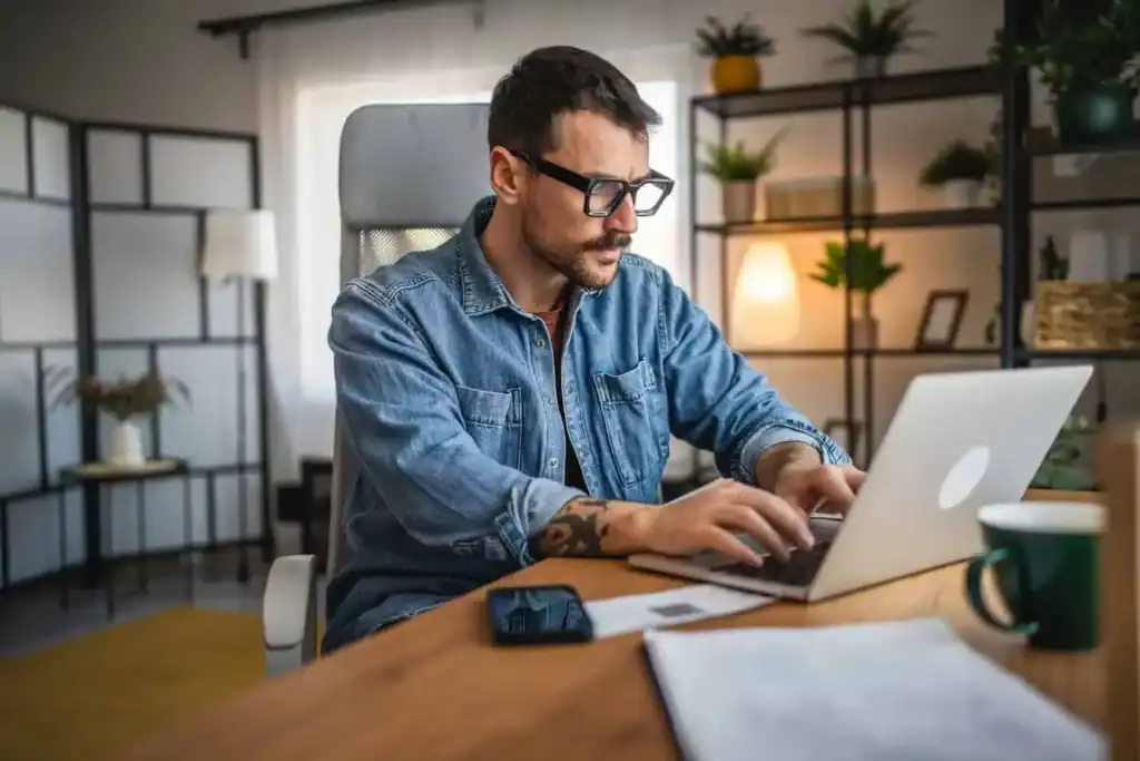 A man in glasses works on a laptop at a wooden desk, researching and planning for a move.