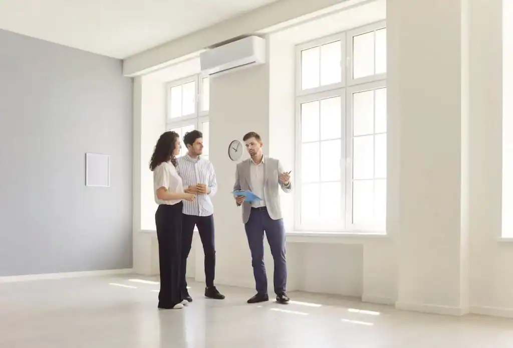 A couple listens to a real estate agent while touring a bright, empty apartment with large windows.