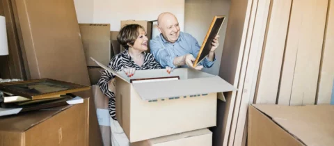 Older couple sorting framed photos and keepsakes while packing boxes during a downsizing move