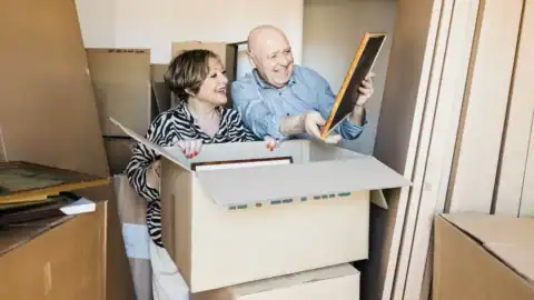 Older couple sorting framed photos and keepsakes while packing boxes during a downsizing move