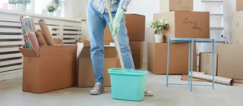 Person mopping the floor in a room filled with moving boxes during a move-out cleaning.