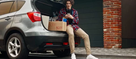 an image of a young man sitting in the open trunk of an SUV on his phone. There are moving boxes visible sitting next to him.
