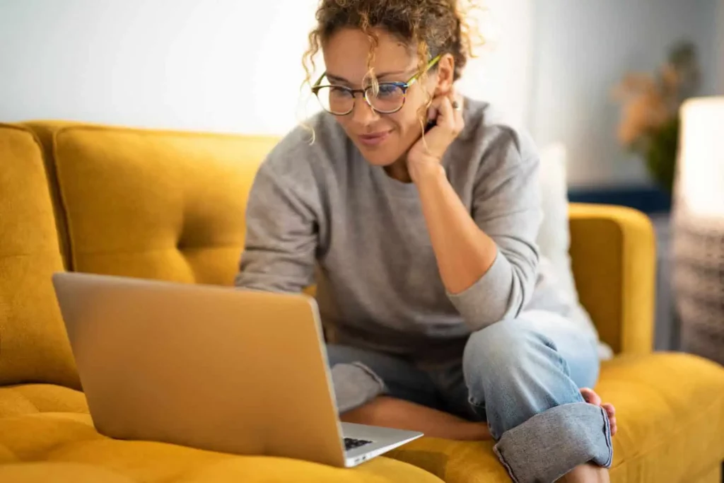 a young woman wearing glasses sits on a yellow couch and researches something on her laptop
