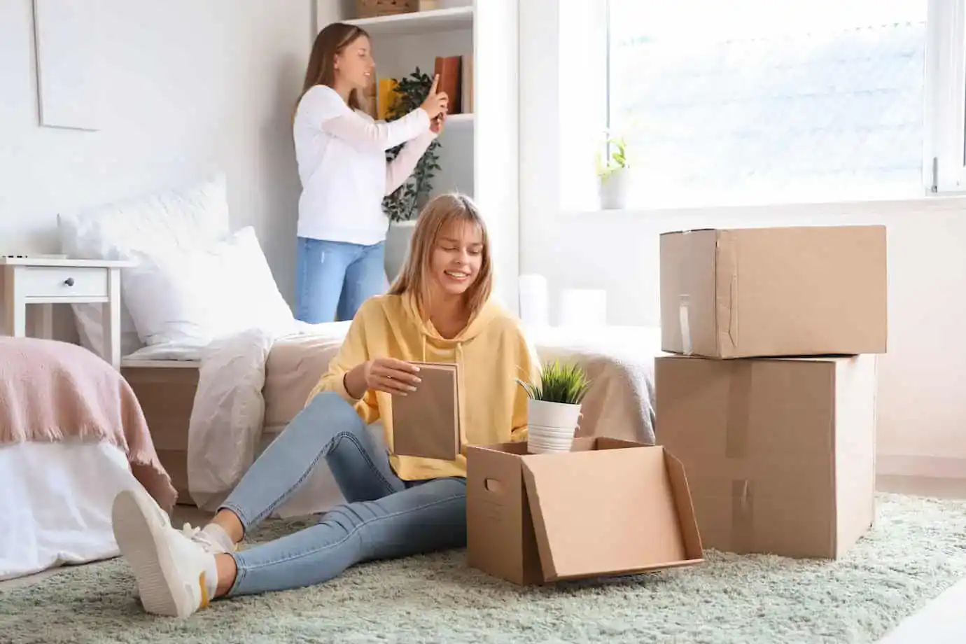 Student unpacking boxes in a college dorm while her roommate arranges items on a shelf in their new dorm