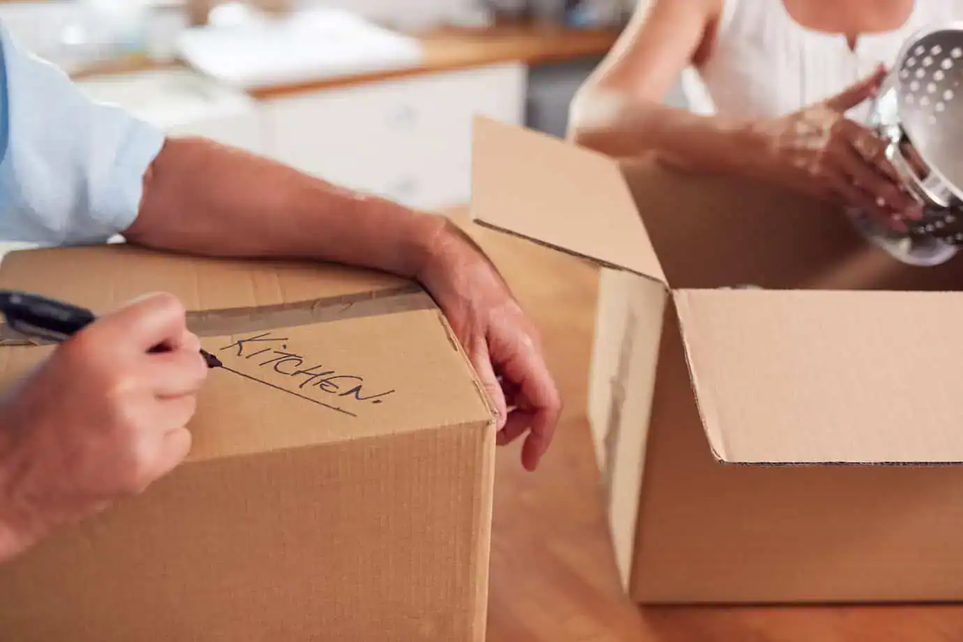 Person labeling a moving box ‘Kitchen’ while packing household items in a kitchen