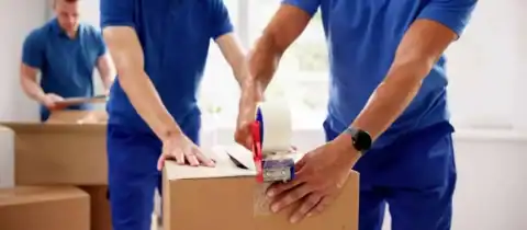 Professional movers sealing a cardboard box with packing tape while preparing items for a move